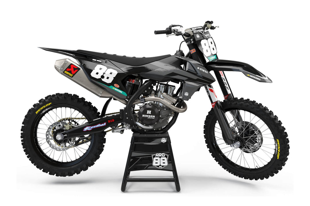 KTM-crosshoj med dekalkitet Black on Grey – svart och grå grafik med breda penseldrag, stora vita siffror och ett turkost band längst ner